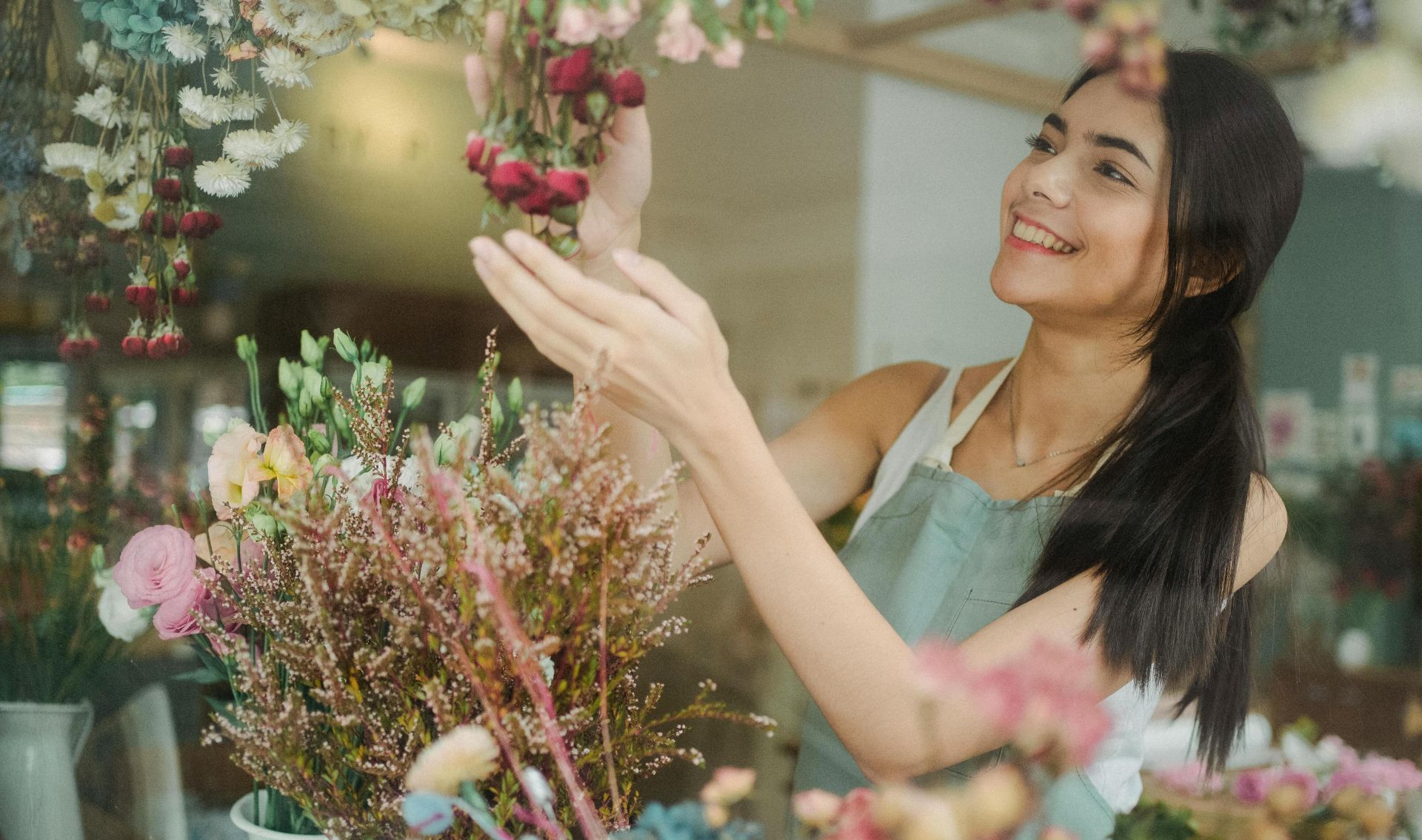 Smiling female florist in apron standing near table with flowers and making bouquets in floral shop