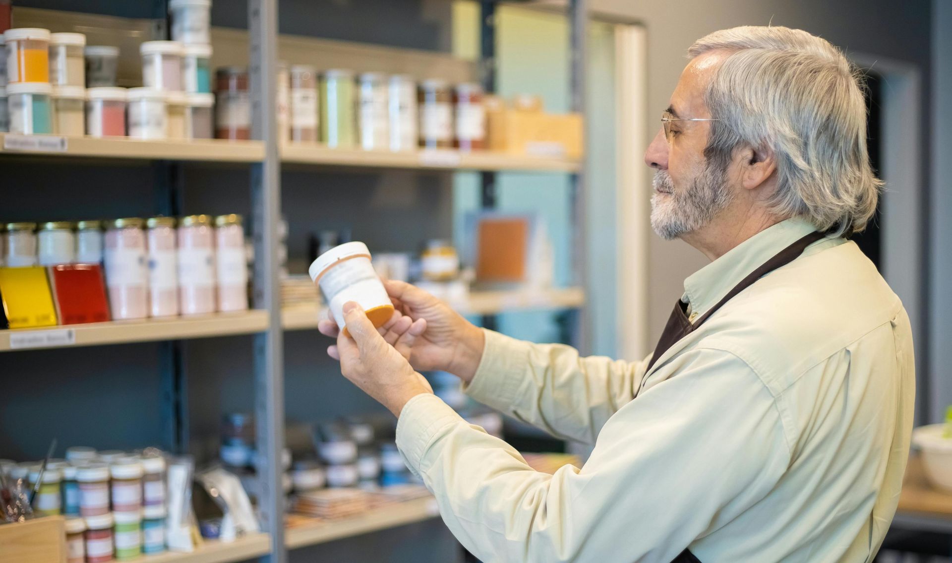 Senior man in apron organizing paint jars on a store shelf indoors.